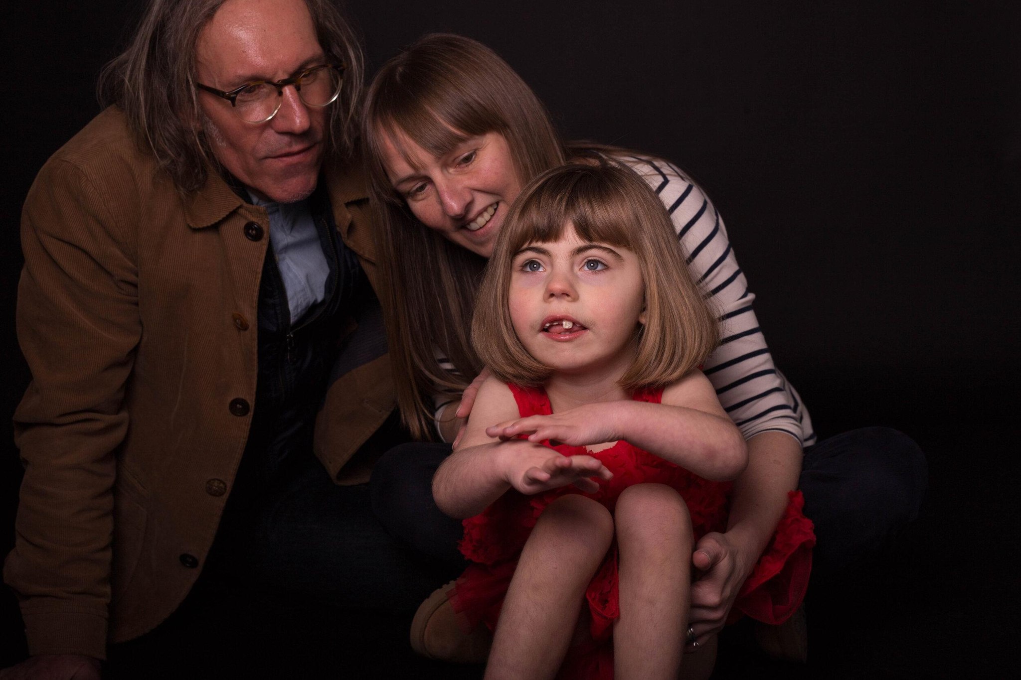 A Maryland family in a dark studio — mother, father, and their daughter in a red dress looking up at the light