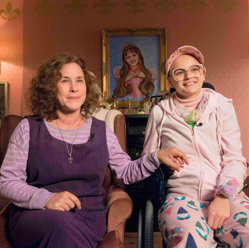 Two women seated together in a pink living room, holding hands and smiling