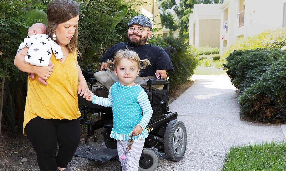 A family on a neighborhood path, father in a power wheelchair, mother carrying a baby, a little girl holding his hand